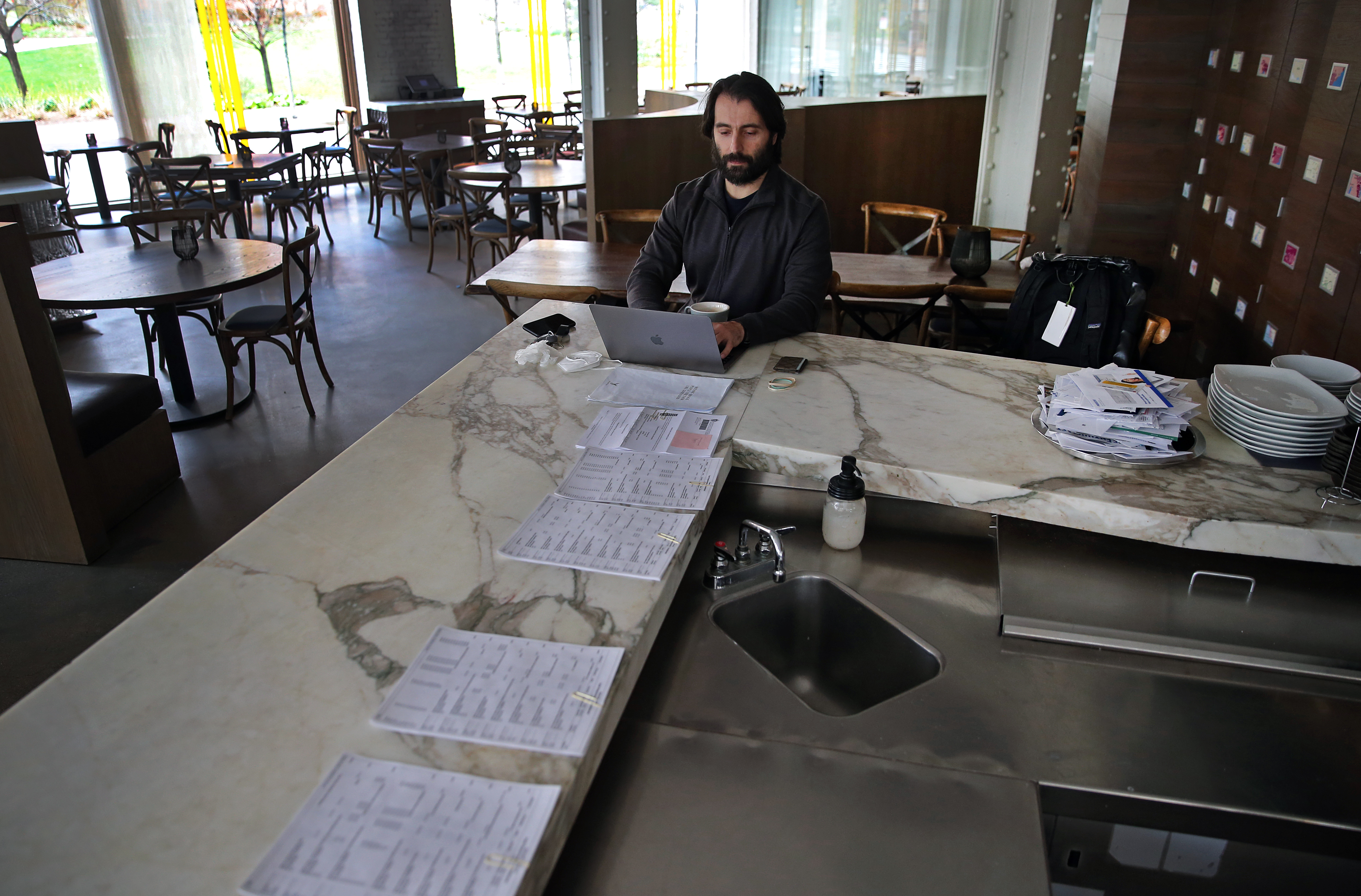 With a row of bills from his restaurants, Eric Papachristos sits at the bar, working on a laptop in his empty Boston restaurant, Trade.