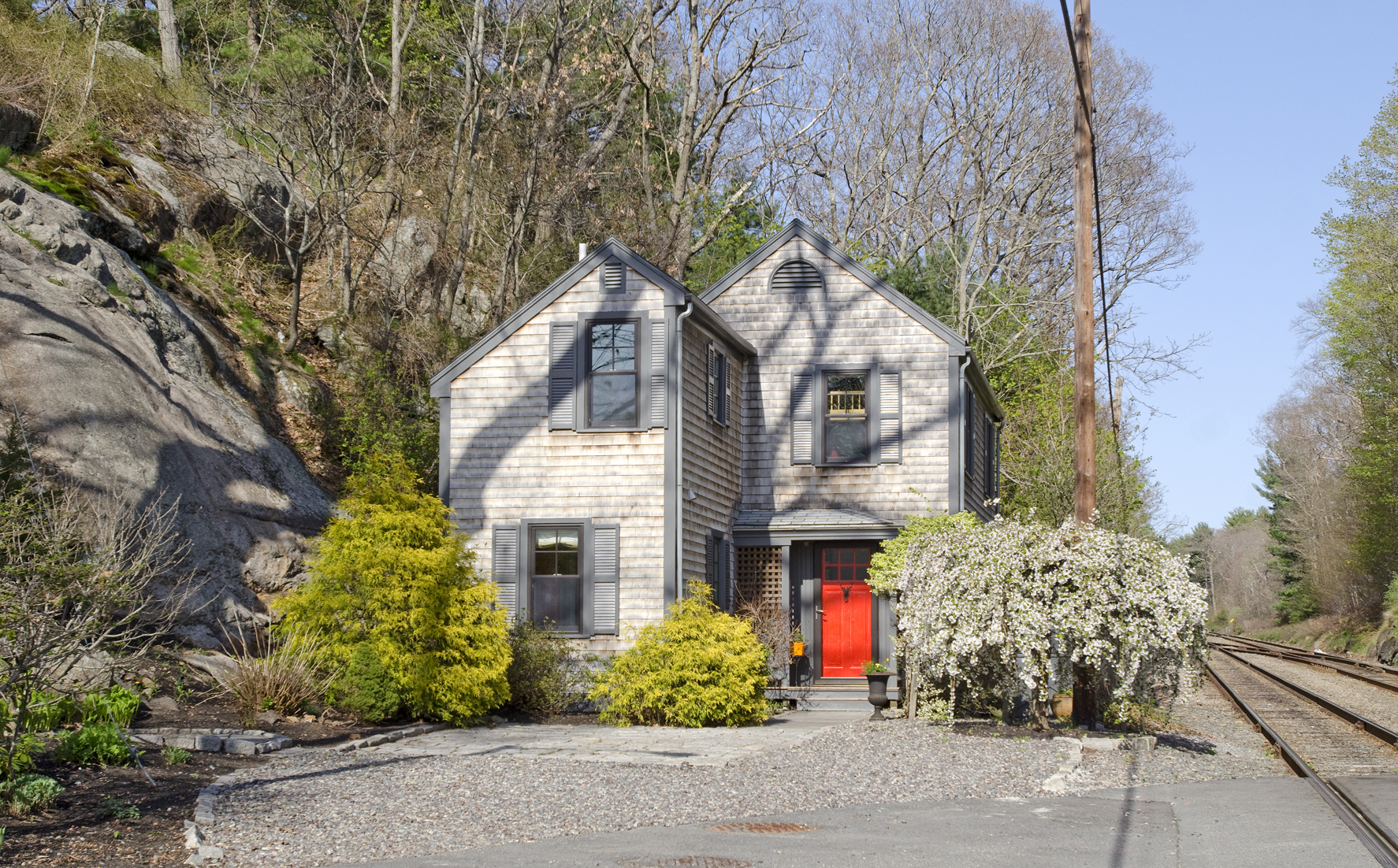 Nestled between a rock ledge and the commuter rail line, the home has a tiny patio and gravel yard. Exterior trim is painted California Paints Gropius Grey.