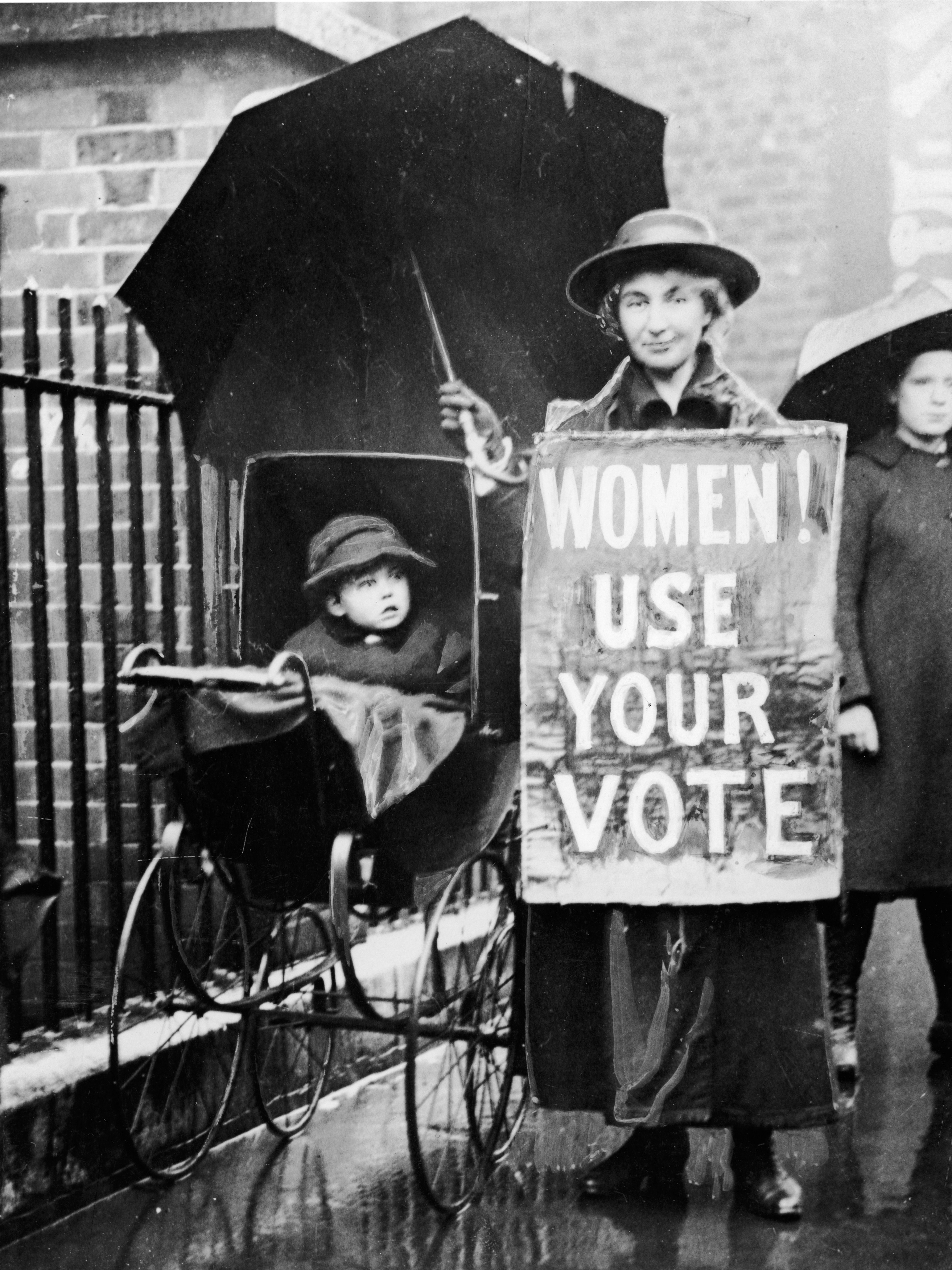 An American suffragette with an umbrella stood next to a baby carriage and wears a sign proclaiming 'Women! Use your vote,' circa 1920.