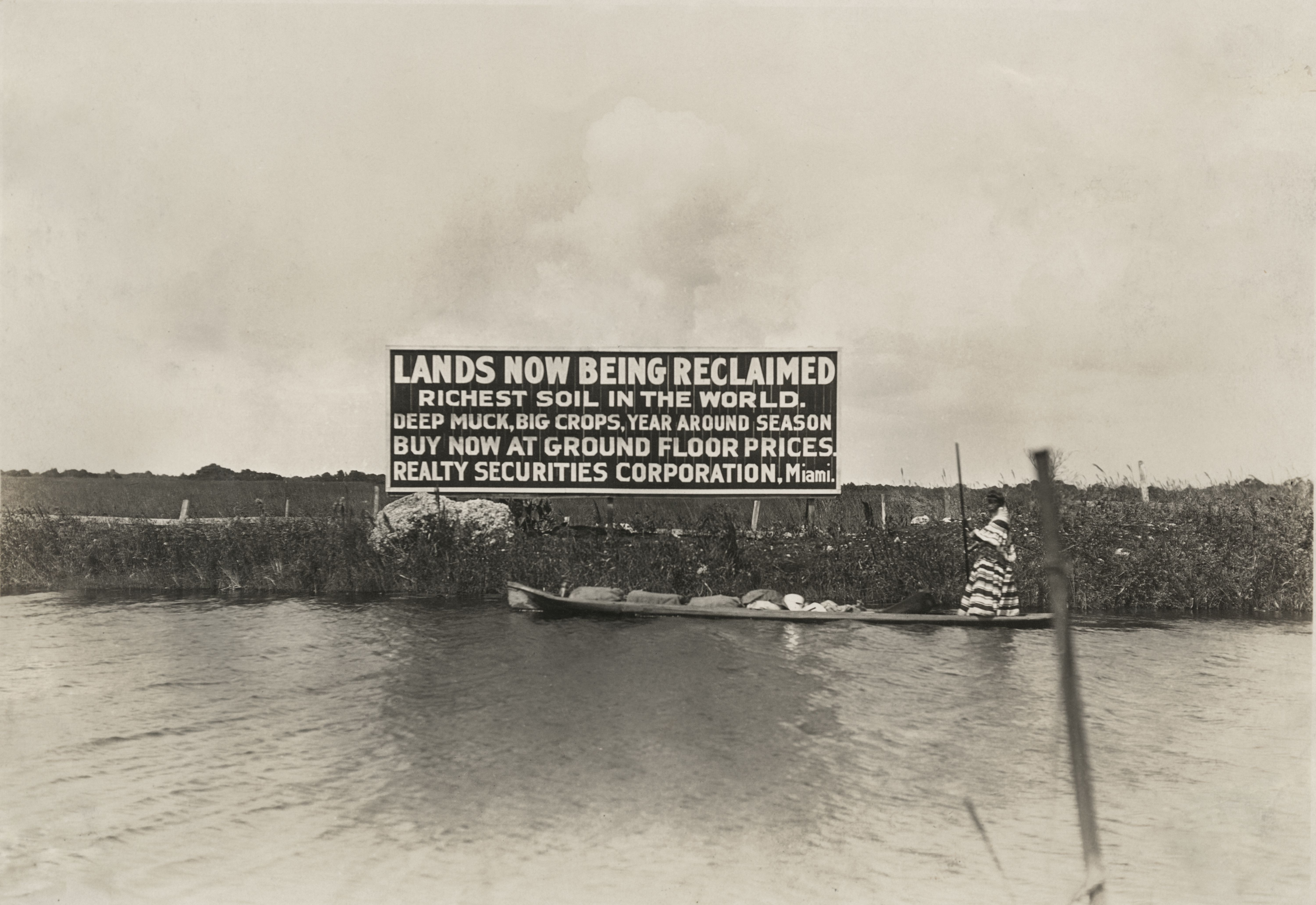 A still image from “The Swamp,” shows a Seminole woman poling past a billboard, advertising the sale of reclaimed Everglades land, circa 1920.