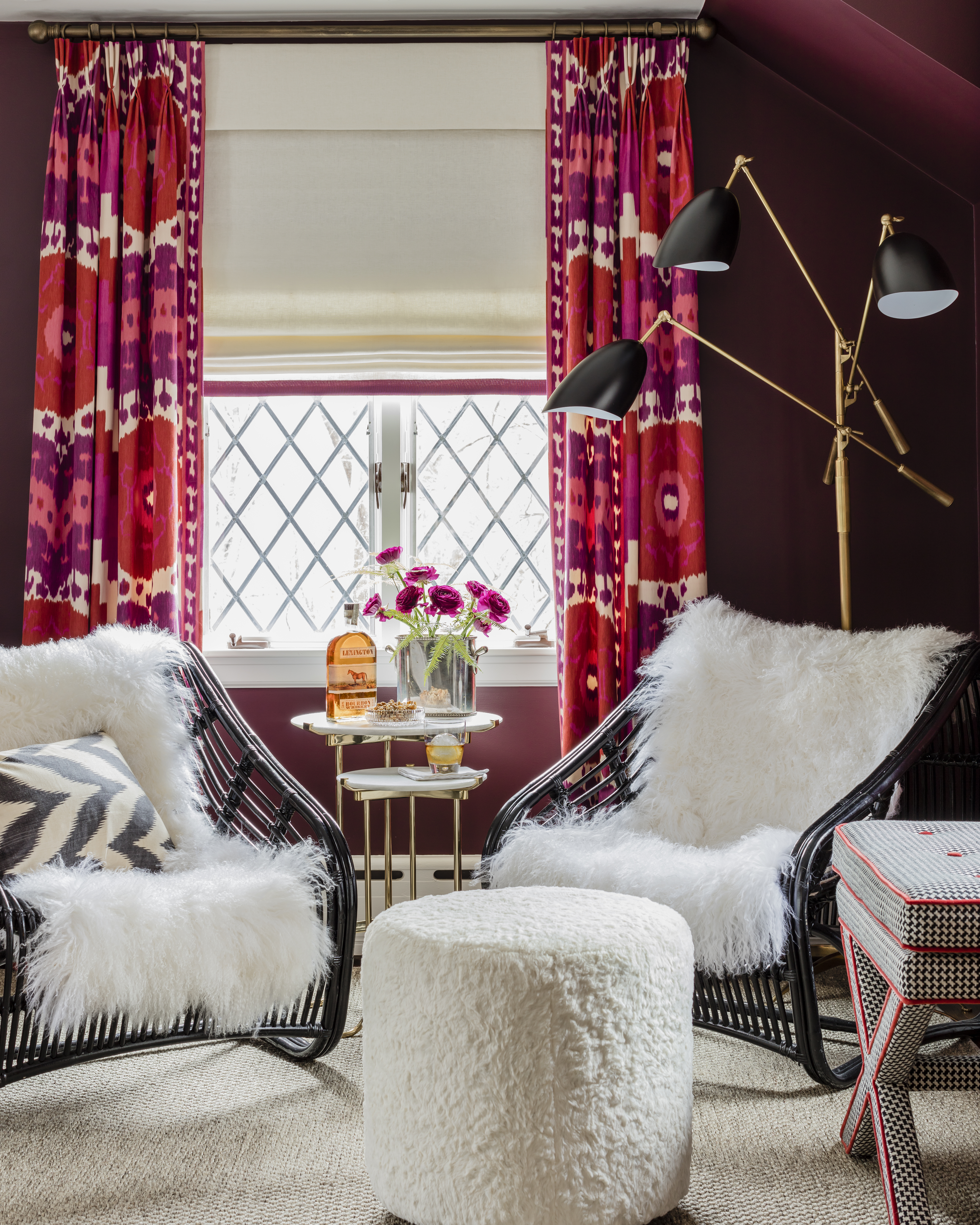 In the sunroom, Gannon installed wow-worthy ikat drapes made of Schumacher fabric; roman shades are cream linen embellished with pink grosgrain ribbon. Tibetan lamb hides accent black rattan chairs. The gold-and-black Visual Comfort floor lamp pairs well with the Shpilners’ bar cart.