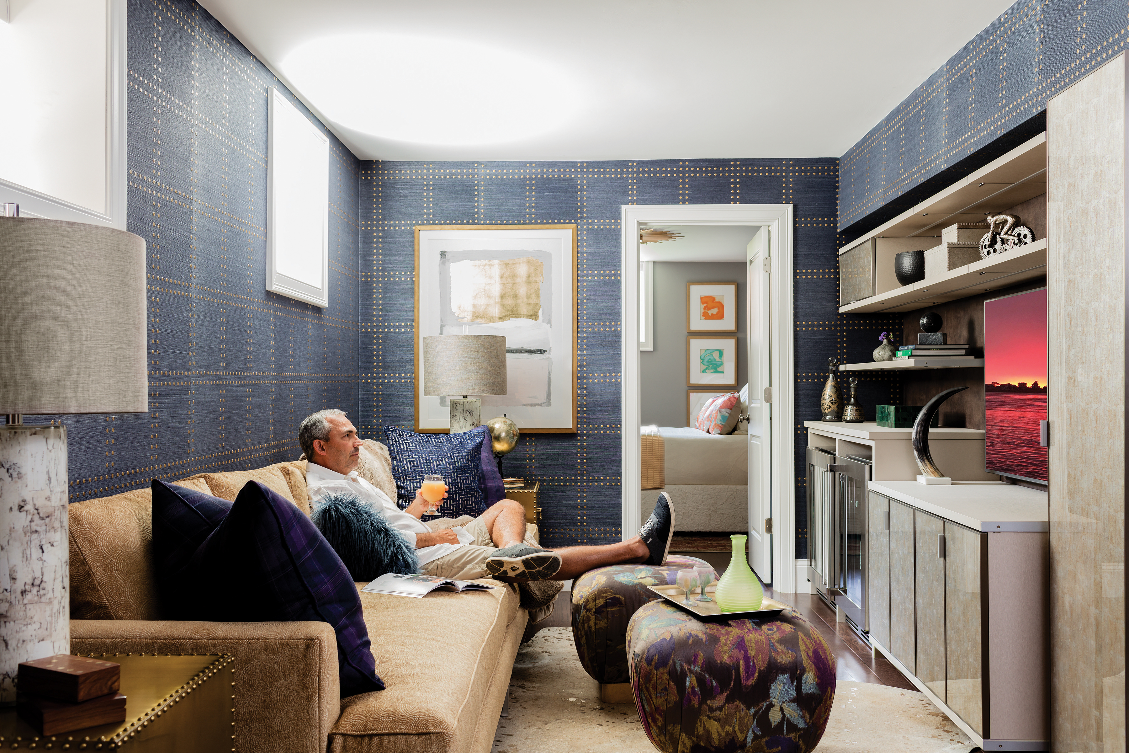 The blue Phillip Jeffries grass-cloth wallcovering is embellished with gold accents that play off the glossy gilt Bernhardt side tables. The custom sofa is by Kravet, and the floral print ottomans, from Moss Studio, serve as both footrests and cocktail tables.