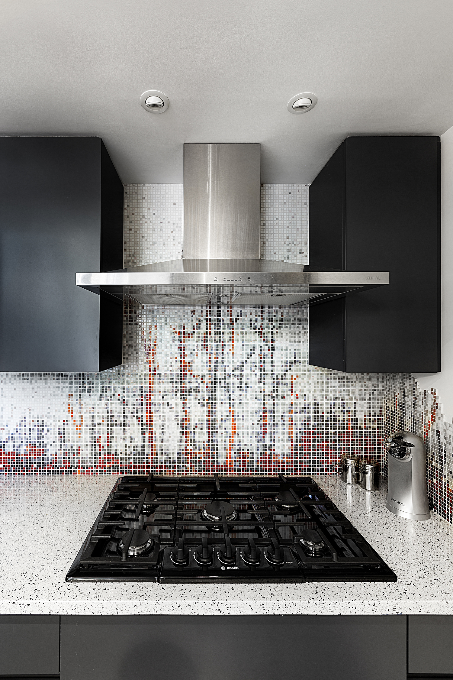 The custom mosaic tile in the kitchen is from Artaic in South Boston. Salem-based North River Design Group built the stained wood cabinetry. A speckled white engineered stone countertop keeps the corner from being too dark.