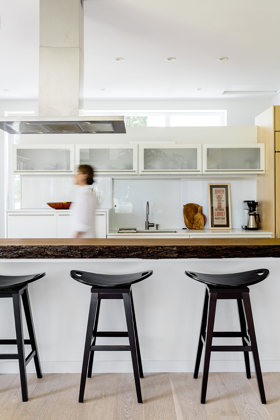 An island topped with a slab of live-edge black walnut and Frampton Saddle bar stools from Wayfair add contrast to the streamlined kitchen area. The freestanding back wall lets light flow while hiding a recycling station and pantry.