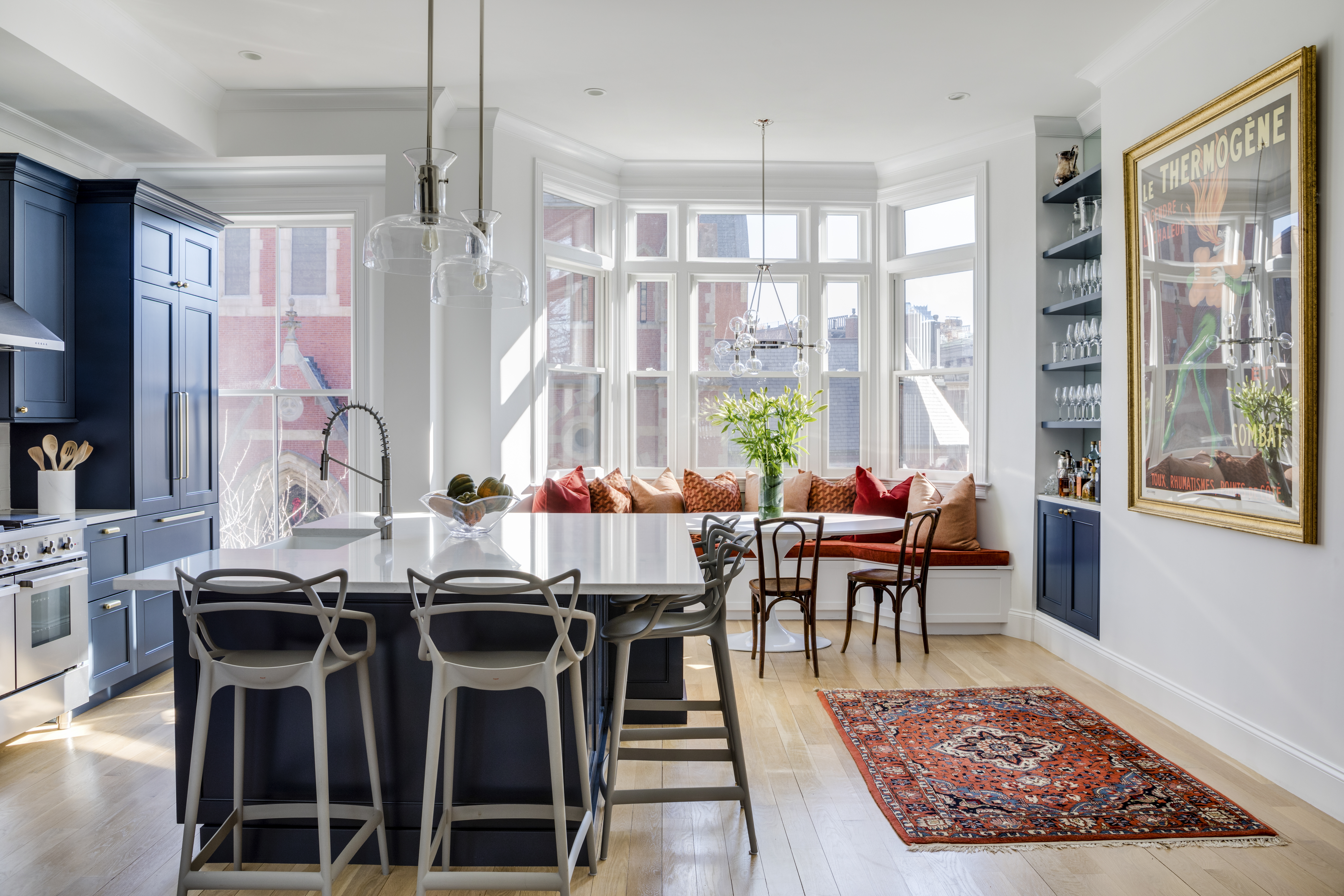 AFTER: The bay window in the breakfast nook overlooks the street. The funky Kartell Masters counter stools echo the lines of the vintage wooden bistro chairs opposite the banquette. Sculptural lights in clear glass — Hudson Valley Lighting Coffey pendants over the island, and a Garim chandelier from Kichler over the table — don’t interfere with the decor or the view.