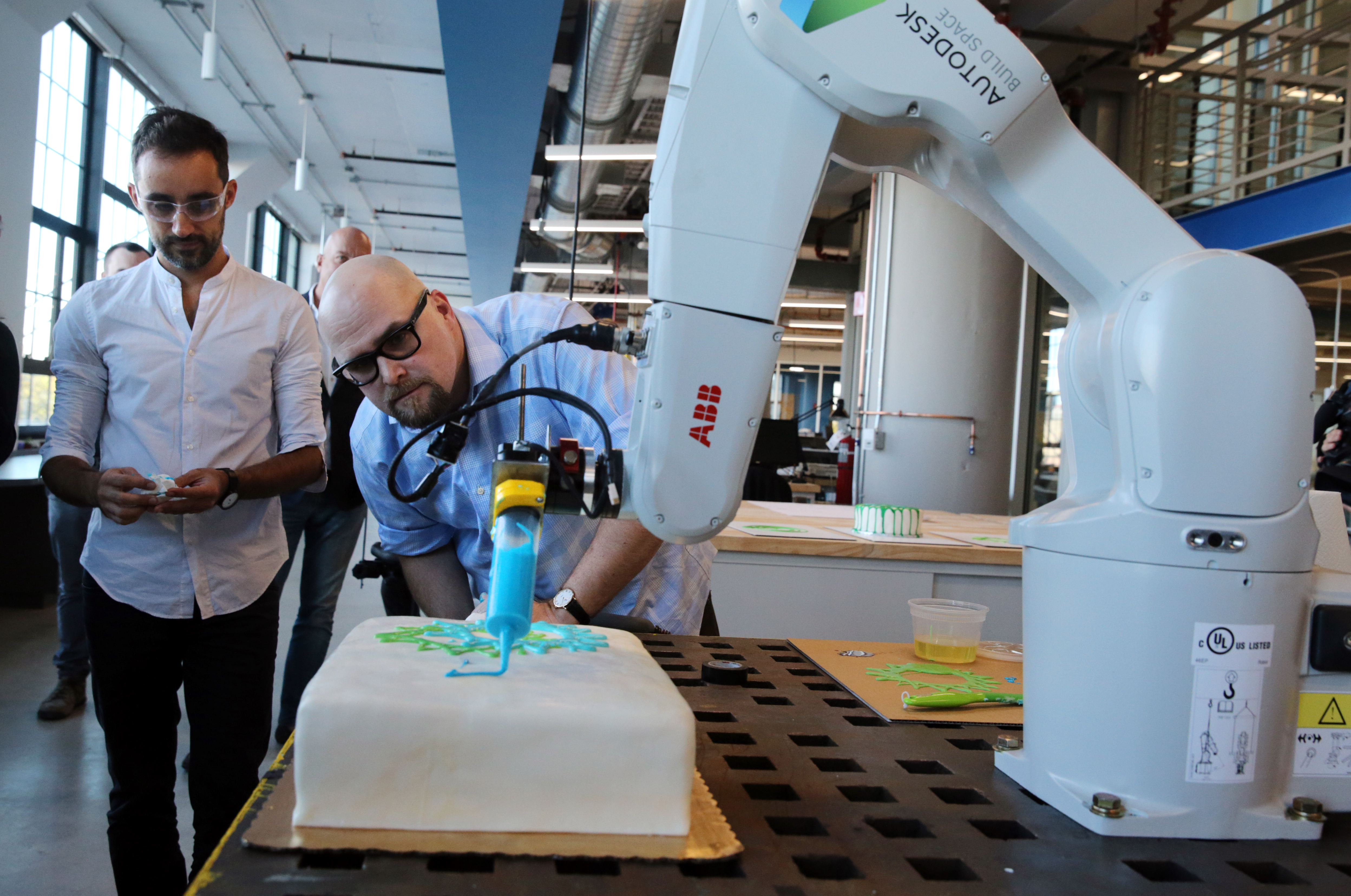 Jose Garcia Del Castillo (far left) and Michael Dewberry use a robotic arm to decorate a cake during a demonstration.