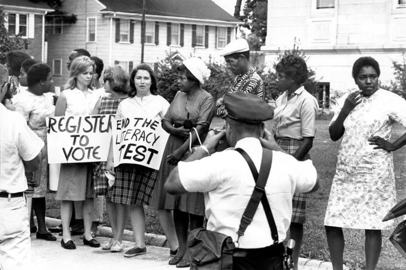 A police officer took a photograph of a group, including Linda Wetmore (plaid dress, holding “Register to Vote” sign), standing outside of the Leflore County Courthouse in Greenwood, Miss., on July 16, 1964.