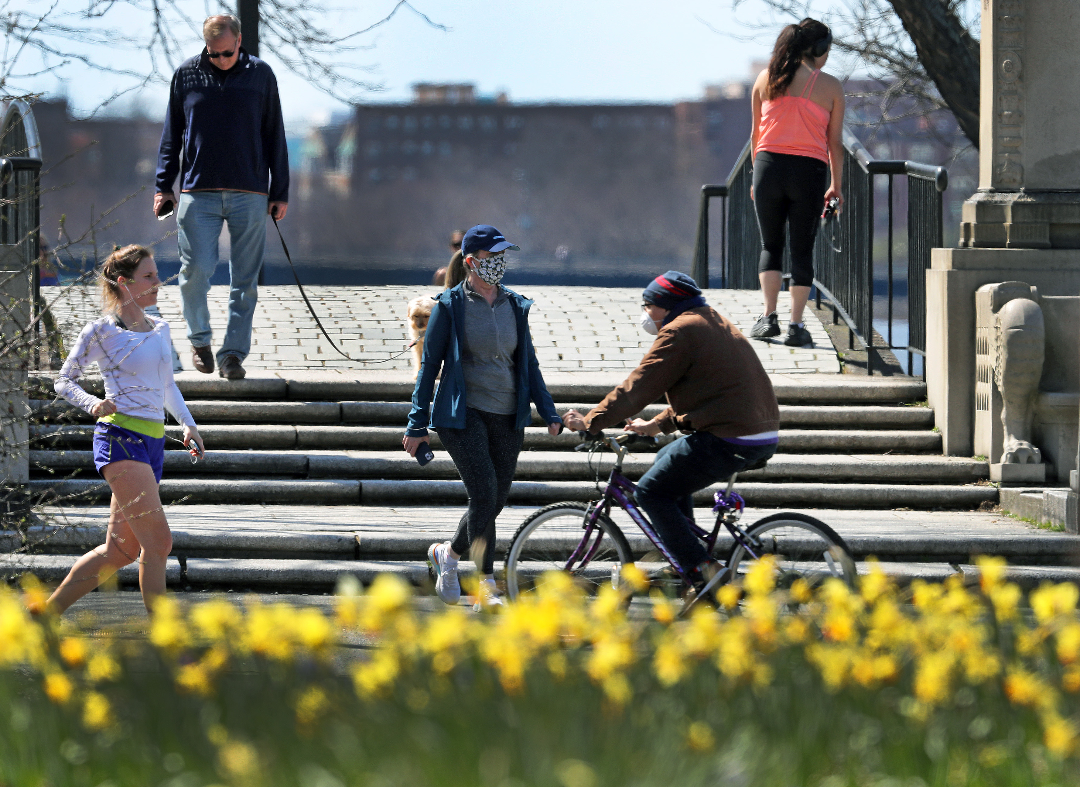 Runners, walkers, a dog, and cyclists, some wearing masks, some not, shared space Monday with some flowers that are in bloom in front of the stage at the Hatch Shell on the Esplanade along the Charles River.